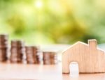 A photo illustration of a wooden carving shaped like a house next to a pile of coins.